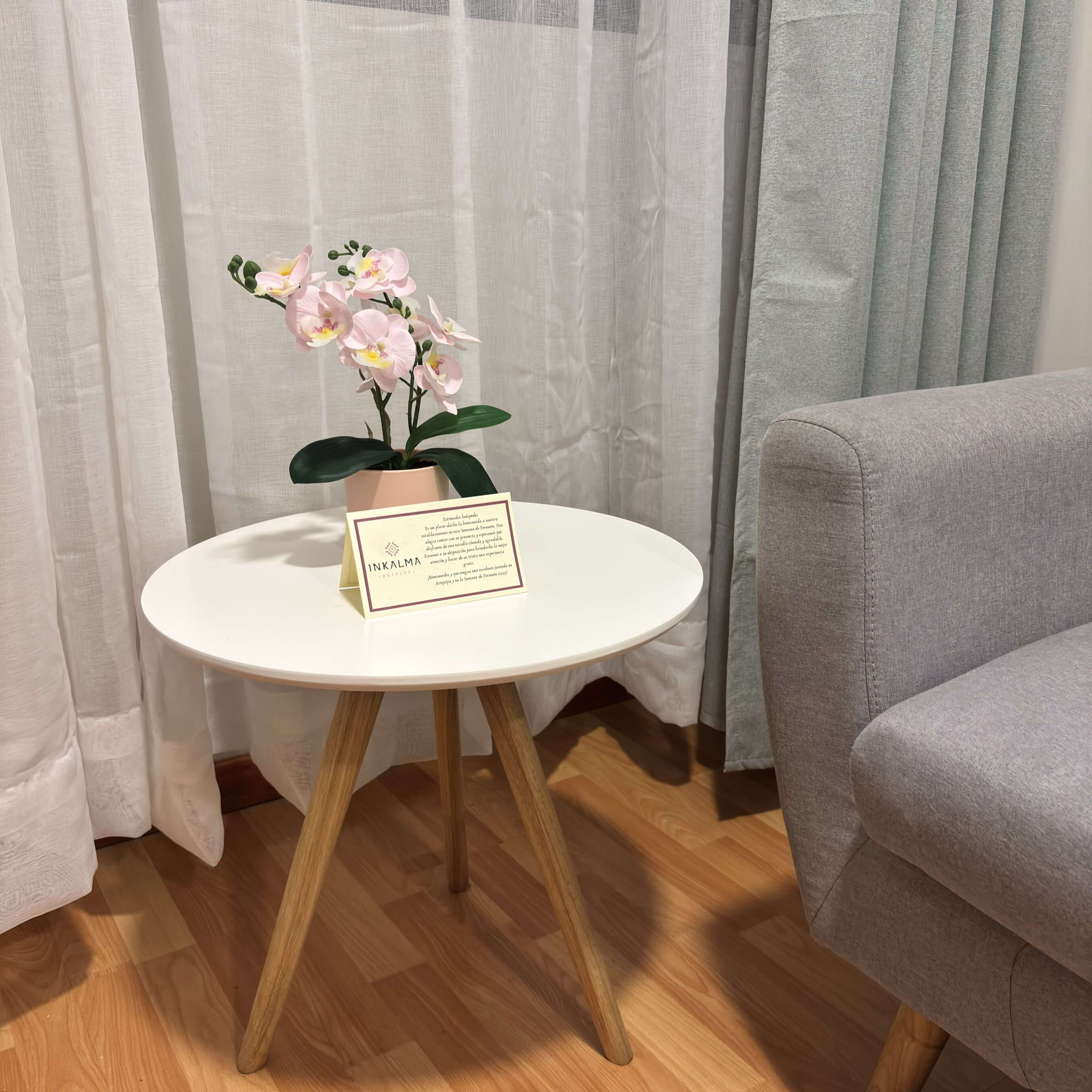 Una pequeña mesa blanca con una orquídea en maceta y una tarjeta, suelo de madera, sofá gris y cortinas de gasa.
