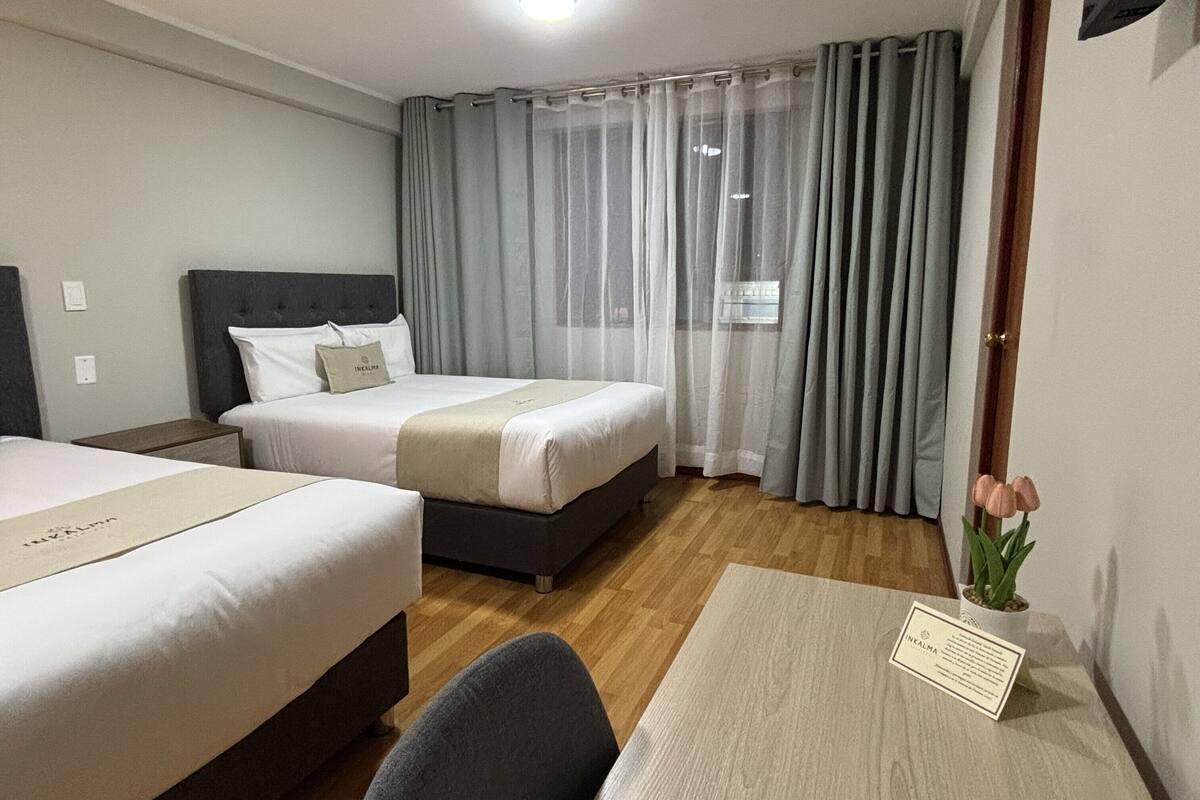 A hotel room with two beds, a desk, a chair, a small table with a card, a potted plant, and a window with curtains.