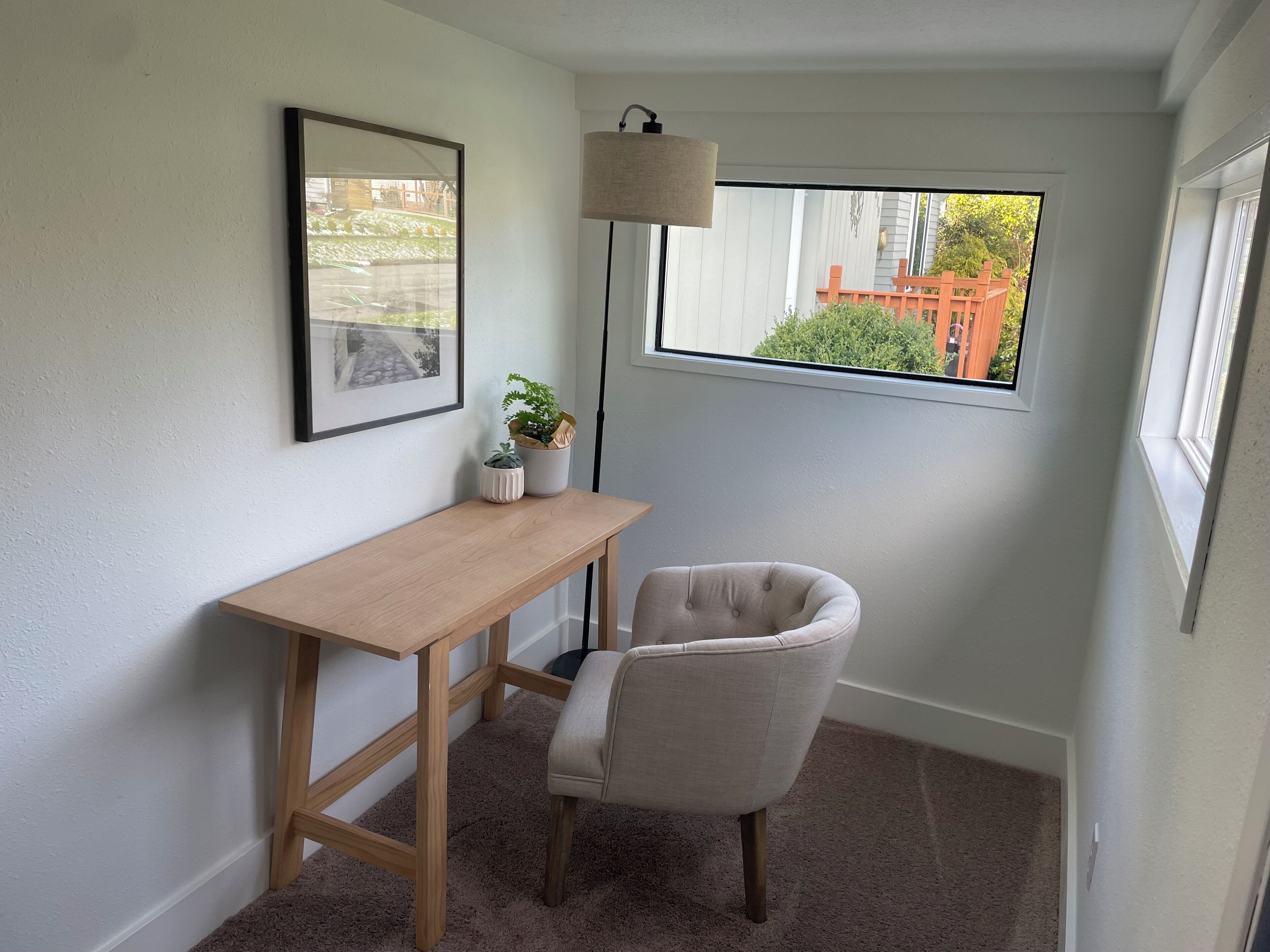 A small, well-lit room with a desk, chair, and a large window offering a view of a red building.