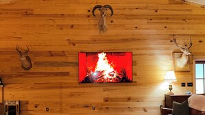 Ett rum med en väggpanel av trä, ett uppstoppat rådjurshuvud på väggen, en öppen spis med en röd låga, en lädersoffa och en matta av kohud.