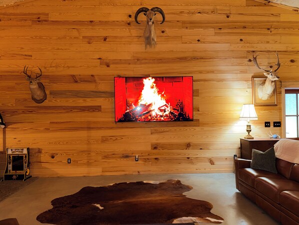 A room with a wooden wall, a mounted deer head, a fireplace with a red fire, a leather couch, and a cowhide rug.