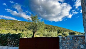 A gated entrance with a stone wall and a metal gate, set against a backdrop of a hilly landscape and a clear blue sky.