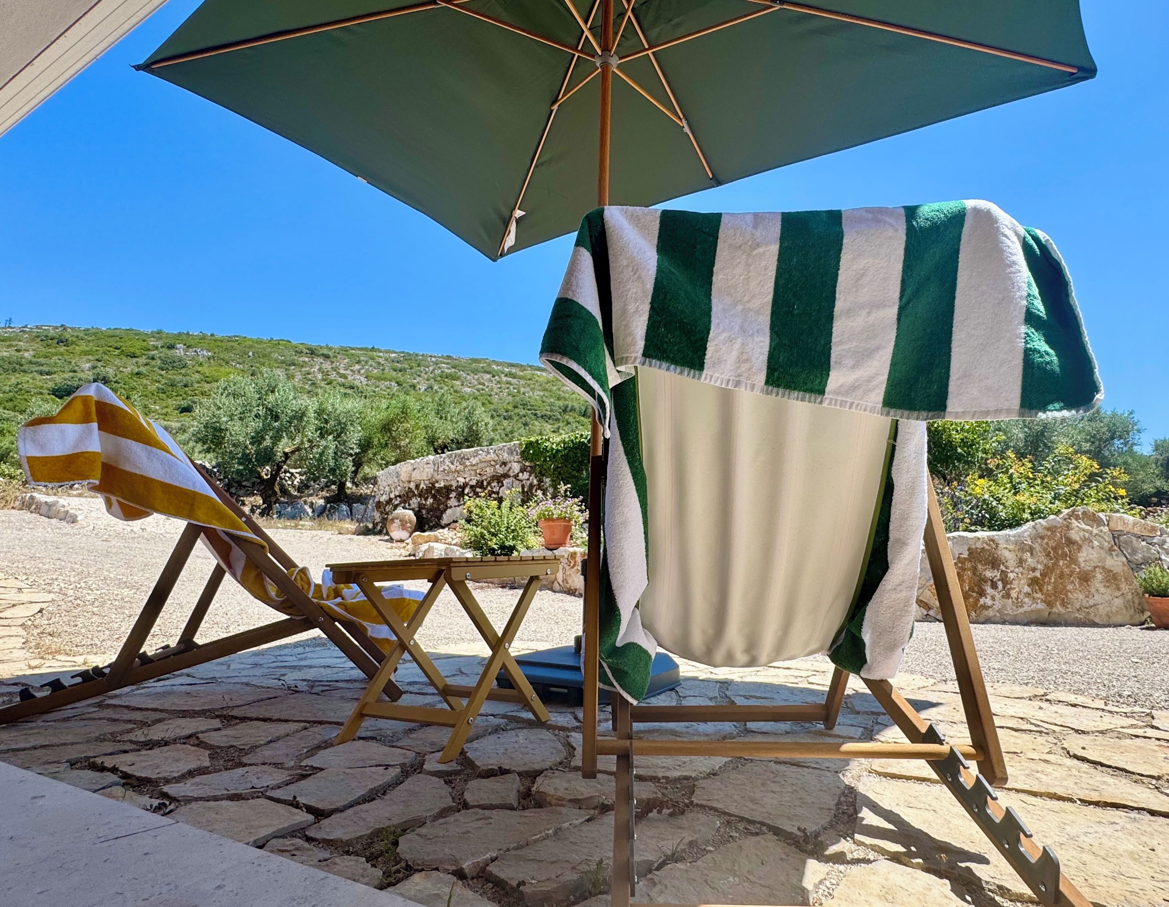 A beachside setting with a wooden chair under a striped umbrella, a stone pathway, and a view of the sea and hills.