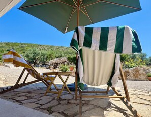 A beachside setting with a wooden chair under a striped umbrella, a stone pathway, and a view of the sea and hills.