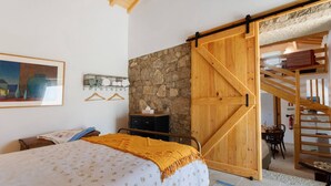 A bedroom with a bed, a wooden wardrobe, a stone wall, a painting, and a staircase.