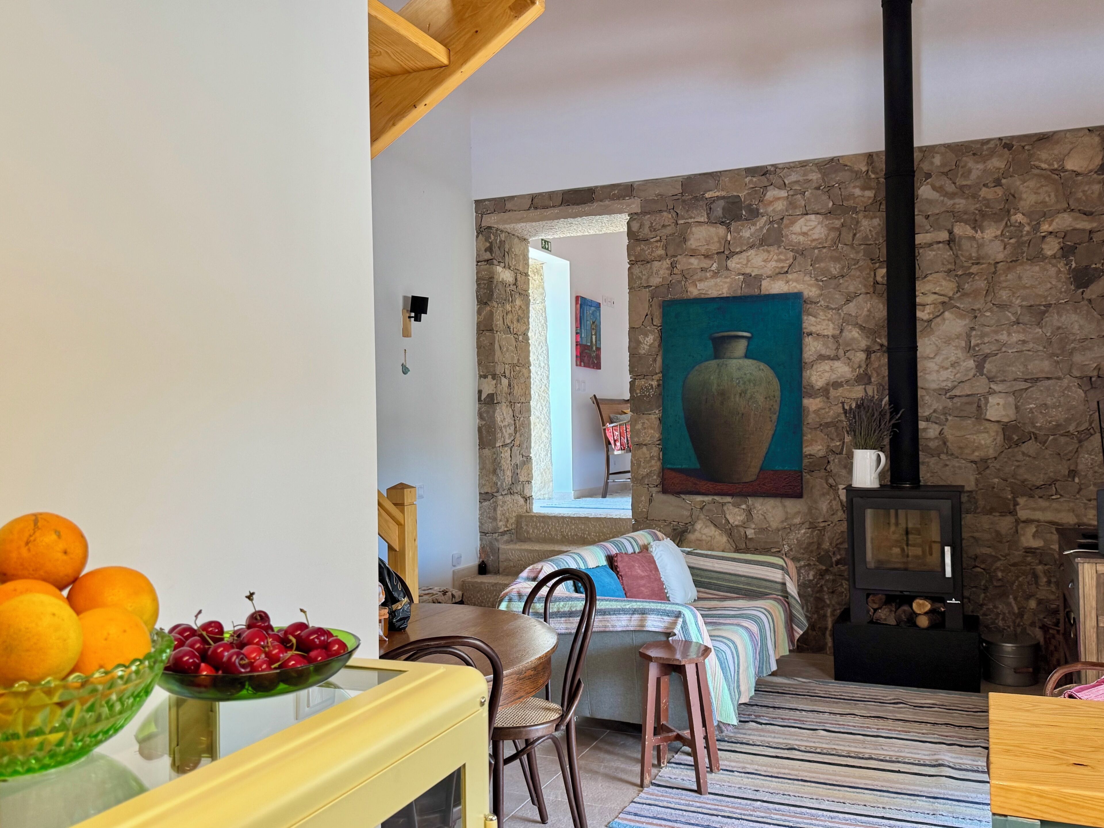 A cozy living room with a stone wall, a painting, a fireplace, and a dining area with fruit bowls.