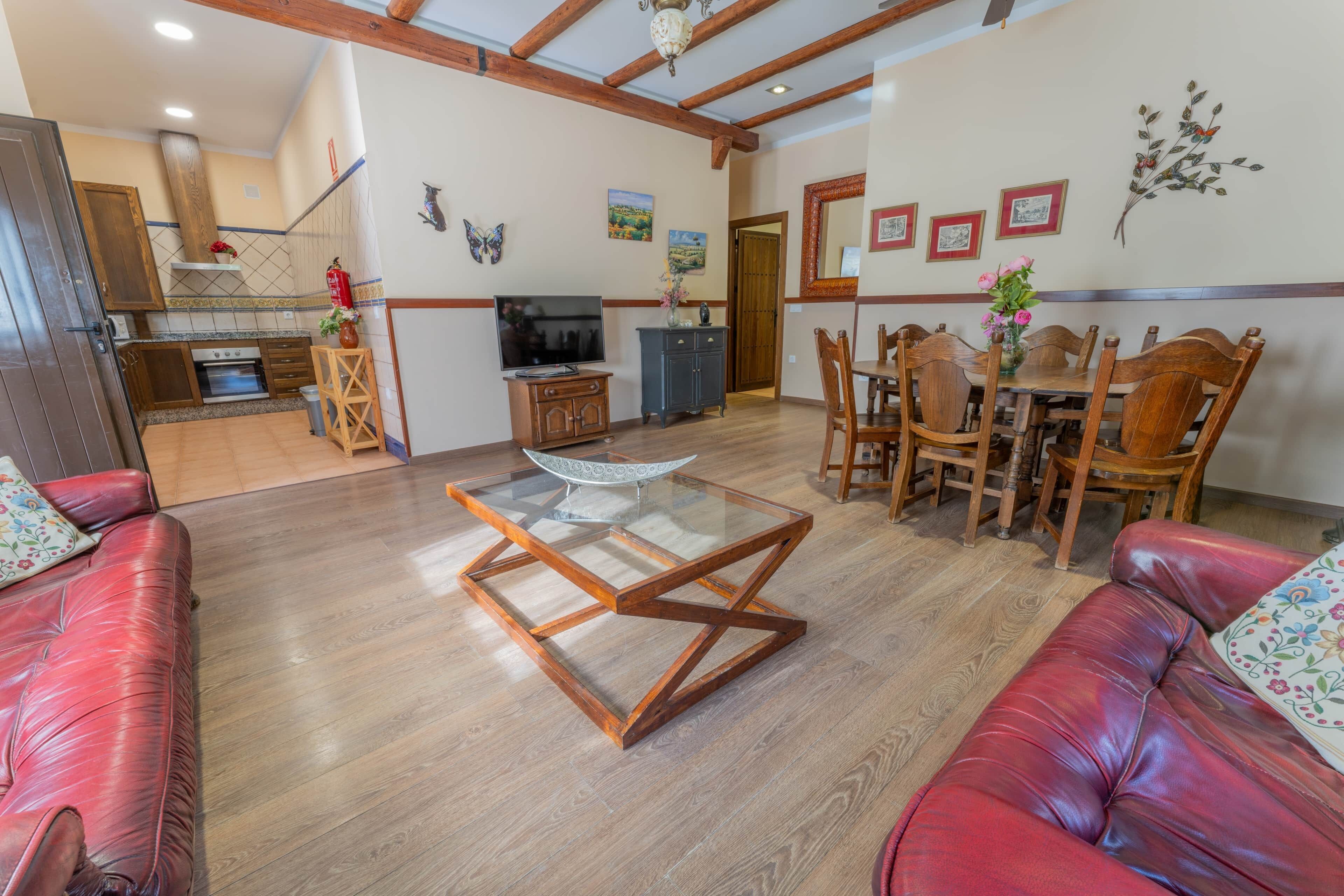 A living room with a red leather sofa, a glass coffee table, wooden dining table with chairs, a kitchen area with a stove, and a TV.