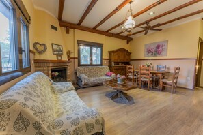 Ein Wohnzimmer mit Kamin, Holzbalkendecke, einer Couch, einem Esstisch mit Stühlen und einer Deckenleuchte.