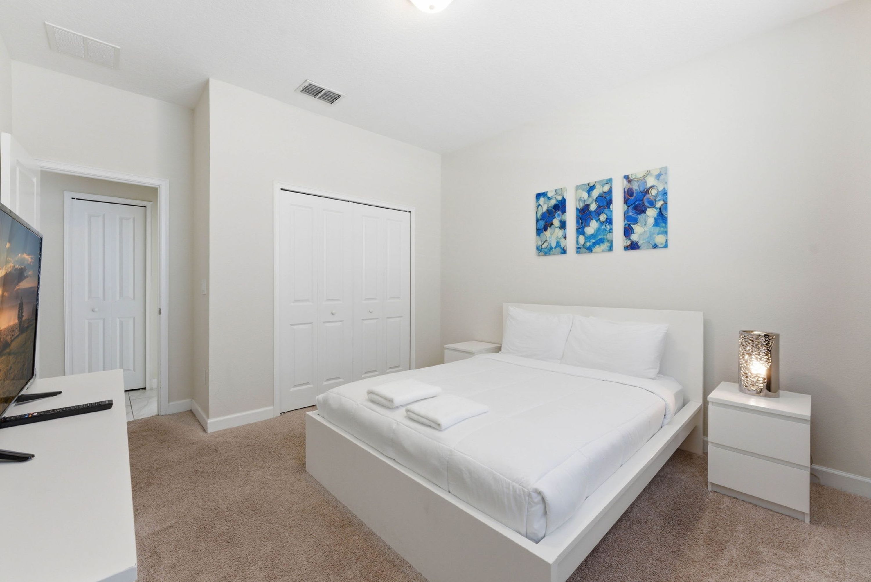 A bedroom with a large bed, white nightstands, a flat-screen TV, and a door leading to a closet.