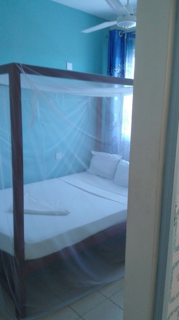 Una cama individual con estructura de madera y ropa de cama blanca en una habitación con paredes color turquesa y ventilador de techo.