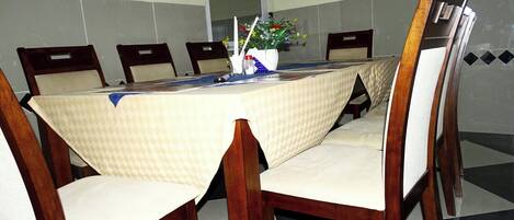 Een vergaderruimte met een tafel, bedekt met een tafelkleed met een patroon, omringd door houten stoelen met beklede zittingen.