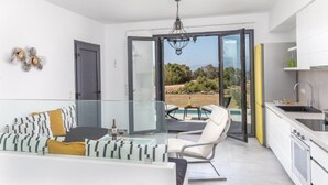 Una sala moderna con un ventanal que da a un balcón, un sofá con cojines estampados, una mesa de comedor con una botella de vino y una cocina con armarios blancos y un lavabo.