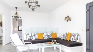 Una sala de estar moderna con una mesa de centro de vidrio, un sillón blanco y un sofá oscuro con cojines estampados.