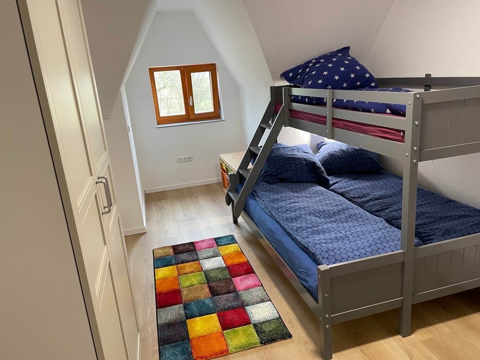 Une chambre avec un lit superposé, une échelle en bois, un tapis coloré et une fenêtre avec des cadres en bois.