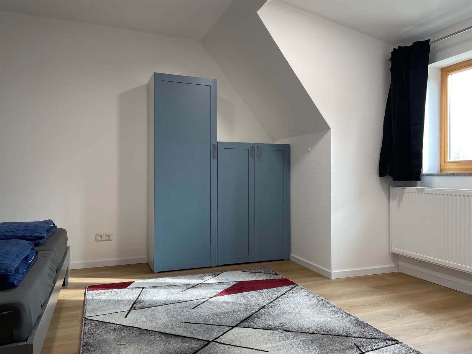 Una habitación con una cama, un armario azul, un tapete geométrico y una ventana con cortinas.