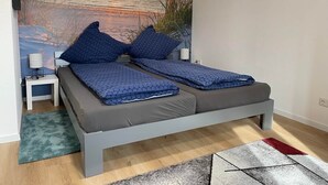 Une chambre à coucher comprenant un lit, une table de chevet, une lampe et une fresque murale représentant un coucher de soleil sur l’eau.