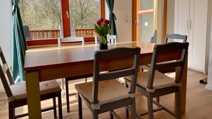 Un coin repas avec une table et des chaises en bois, un vase rempli de fleurs et une vue sur les arbres à travers de grandes fenêtres.