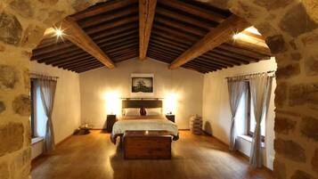 A bedroom with a stone wall, wooden beams, a bed, bedside tables, and a window with curtains.
