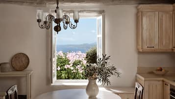 Un comedor con una mesa redonda, un florero con plantas, una araña y una ventana que ofrece vistas a flores rosadas y un paisaje lejano.