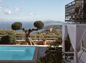 Un espace aménagé au bord de la piscine, offrant une vue sur la mer et les collines, avec une clôture en bois et une chaise blanche dotée d’un parasol.