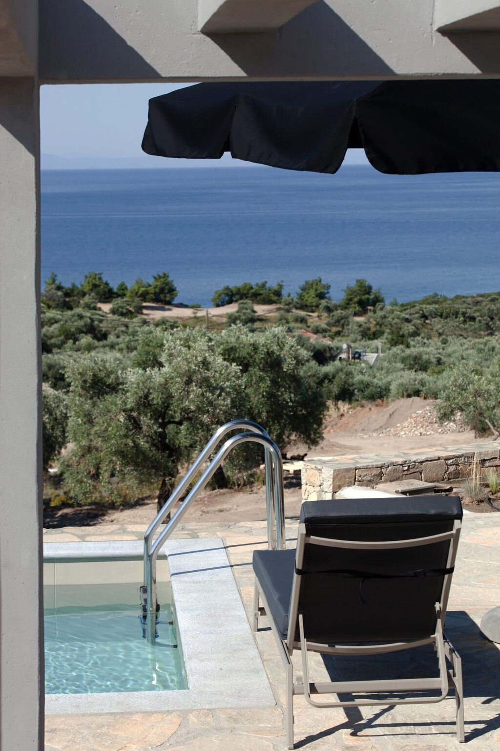 Un espace aménagé au bord de la piscine, avec une chaise et une échelle métallique menant à l’intérieur de la piscine, offrant une vue sur un paysage côtier avec des arbres et la mer.