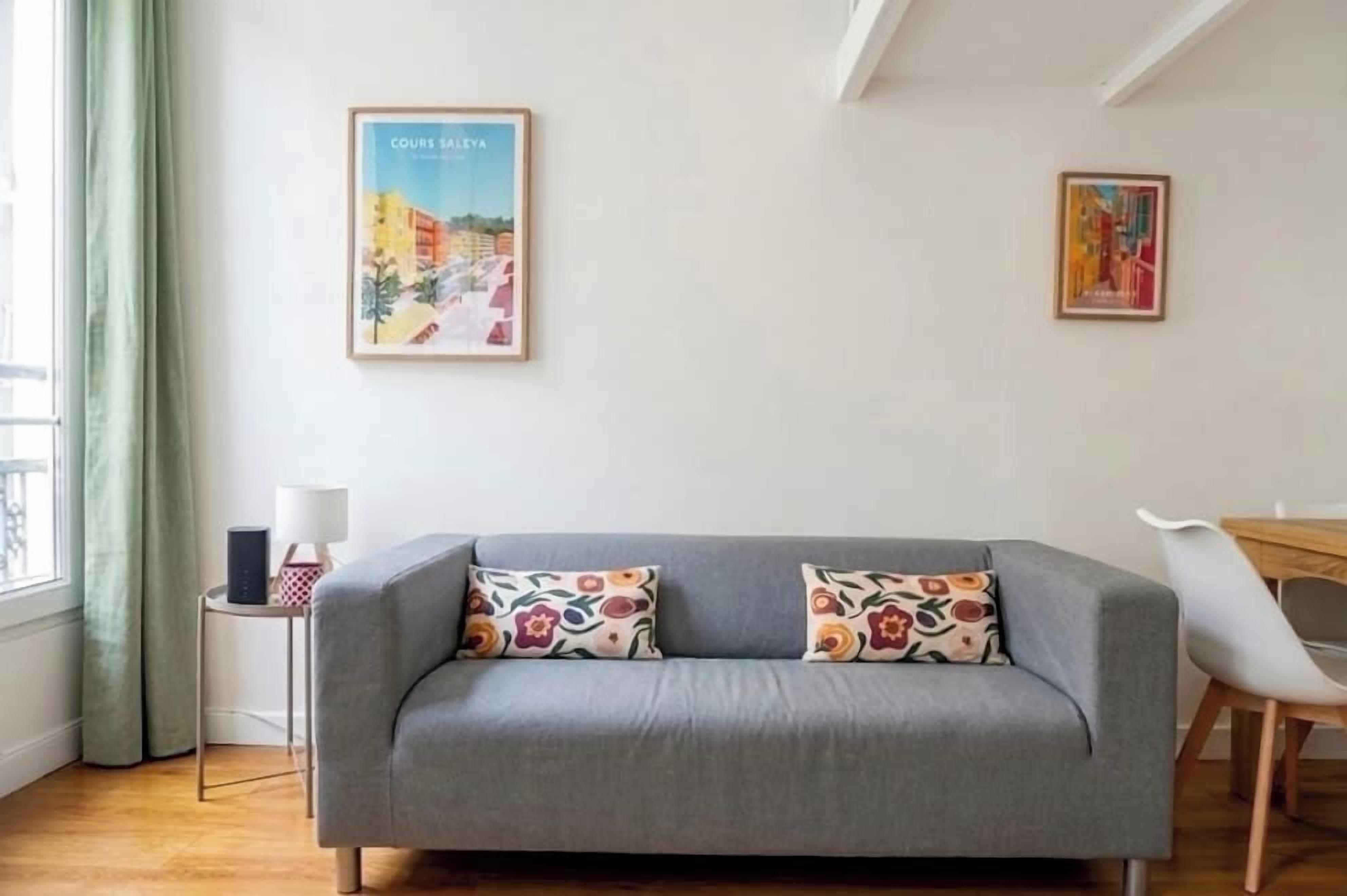 Een moderne woonkamer met een grijze bank, twee gekleurde kussens, een klein bijzettafeltje met een lamp en twee ingelijste kunstwerken aan de muur.