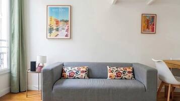 Een moderne woonkamer met een grijze bank, twee gekleurde kussens, een klein bijzettafeltje met een lamp en twee ingelijste kunstwerken aan de muur.