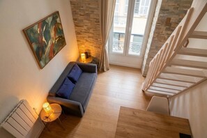 A modern living room with a sofa, a small table with a lamp, a radiator, and a staircase leading to an upper floor. - Le Siagre - 5min à pied de la gare (Nice)
