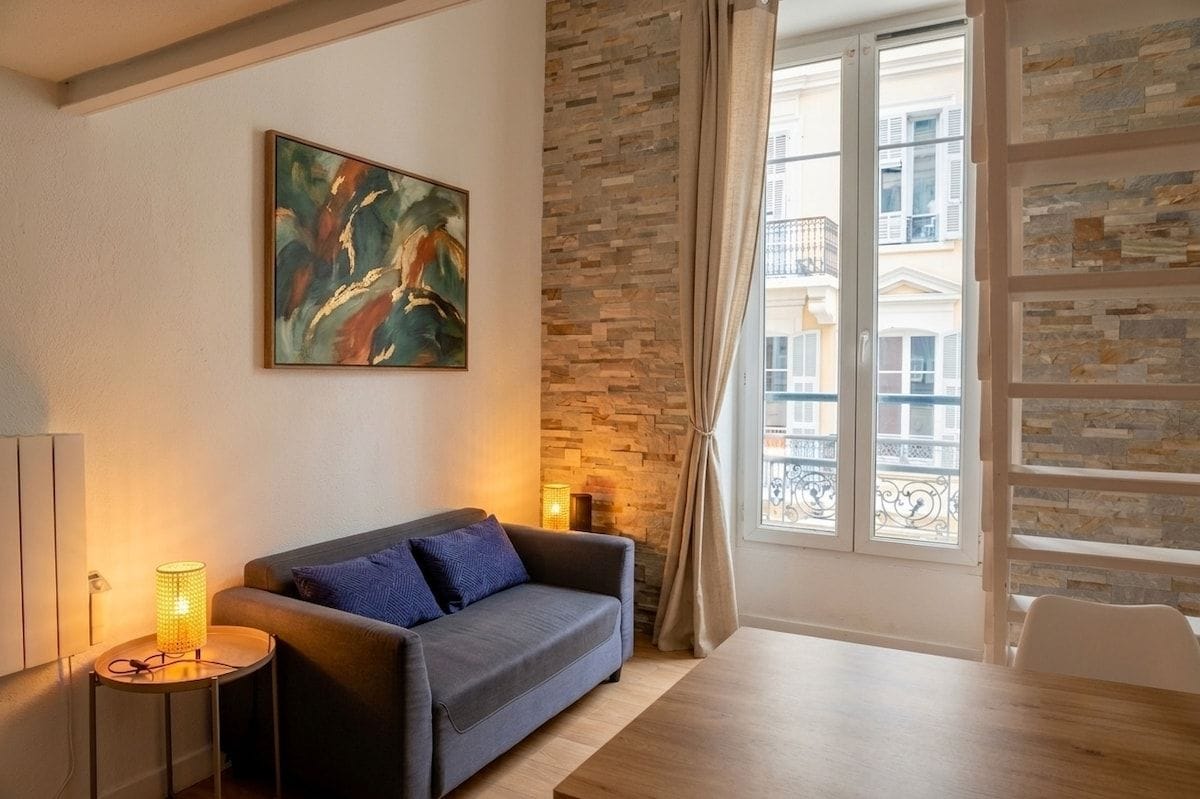 Un salon avec un canapé, un tableau, une lampe, un mur en pierre et une fenêtre donnant sur des immeubles.