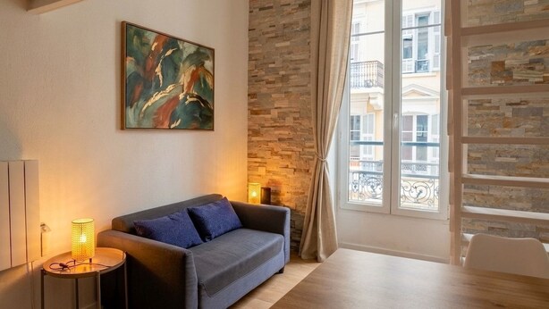 Ett vardagsrum med en soffa, en tavla, en lampa, en vägg av sten och ett fönster med utsikt över byggnader.