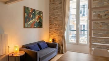 Un salon avec un canapé, un tableau, une lampe, un mur en pierre et une fenêtre donnant sur des immeubles.