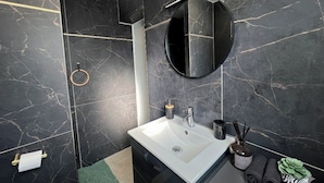 Un baño moderno con un lavamanos blanco, paredes de mármol negro, un espejo redondo y un toallero con toallas dobladas.
