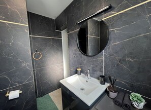 Un baño moderno con un lavamanos blanco, paredes de mármol negro, un espejo redondo y un toallero con toallas dobladas.