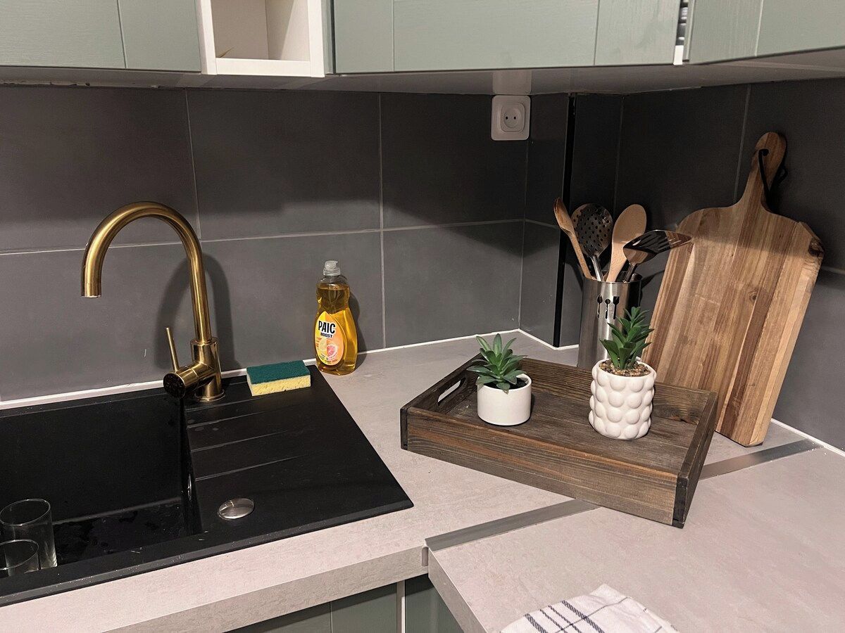 Una cocina con un lavabo negro, un grifo dorado, una botella de detergente, una tabla de cortar de madera y una bandeja con una planta en maceta.