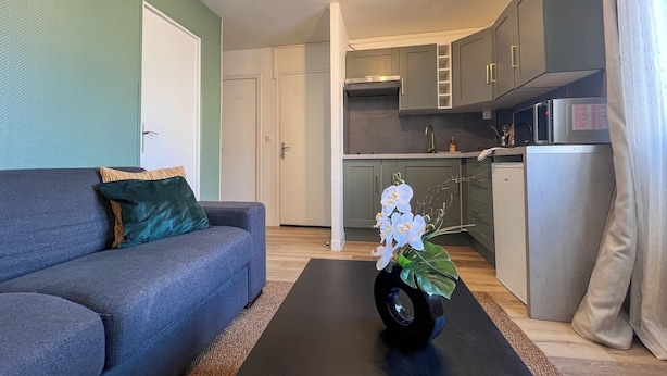 Una sala de estar con un sofá, una mesa de centro con un florero, y una zona de cocina con armarios y electrodomésticos.