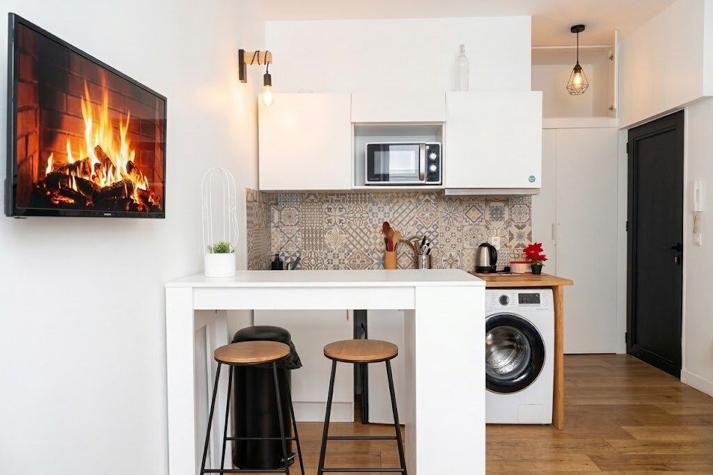 Een kleine keuken met een flatscreen televisie, een magnetron, een wasmachine en twee barkrukken.
