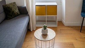Ein Wohnzimmer mit einer Couch, einem kleinen runden Tisch mit einer Pflanze, einem Fenster mit Vorhängen und einem an der Wand befestigten Regal.