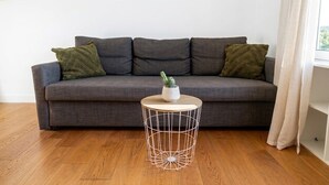 Ein graues Sofa mit drei Kissen, ein runder Tisch aus Drahtgestell mit einer Topfpflanze und ein Holzboden.