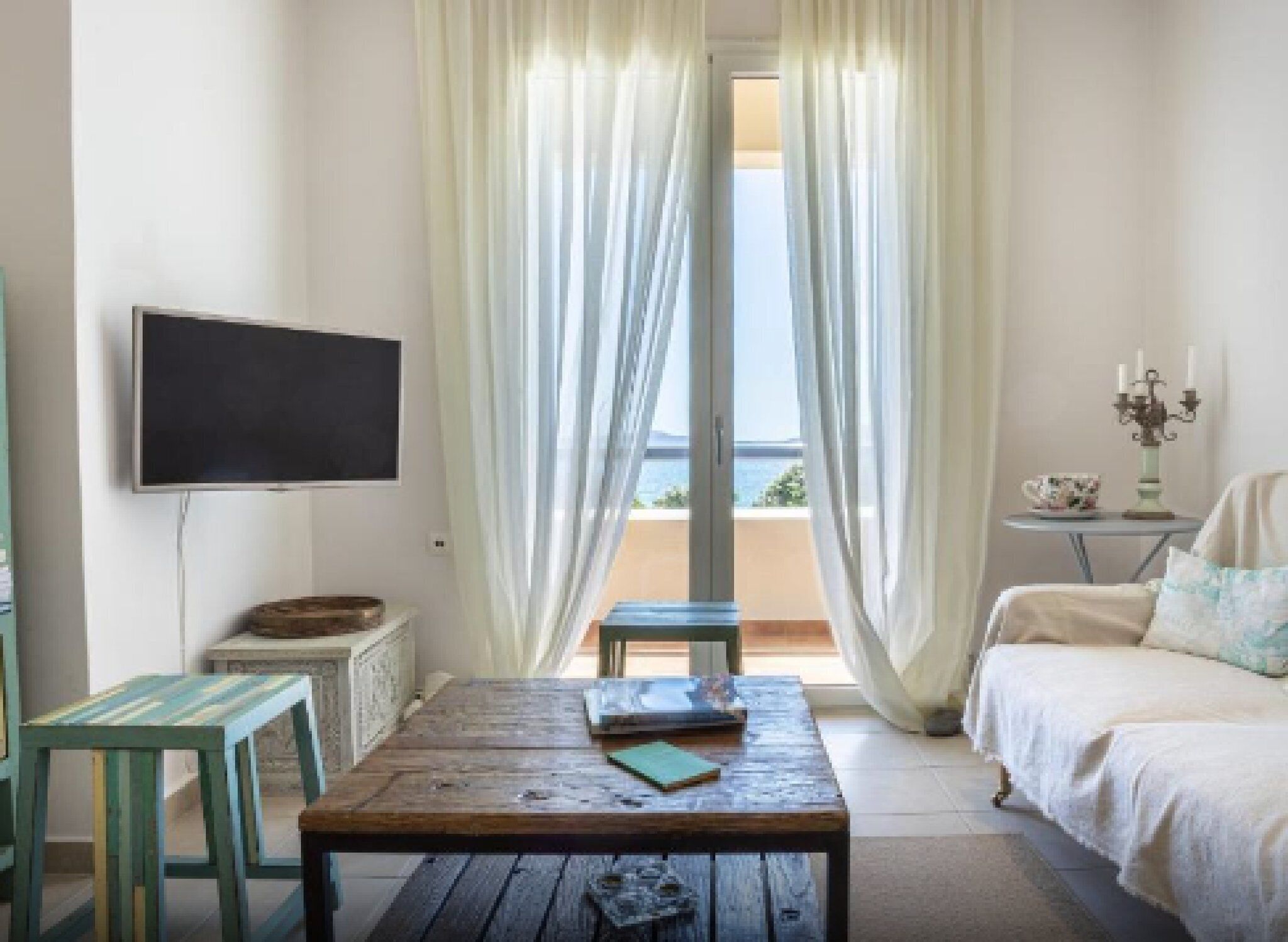 A room with a wooden table, a bed, a TV, and a view of the sea through sheer curtains.