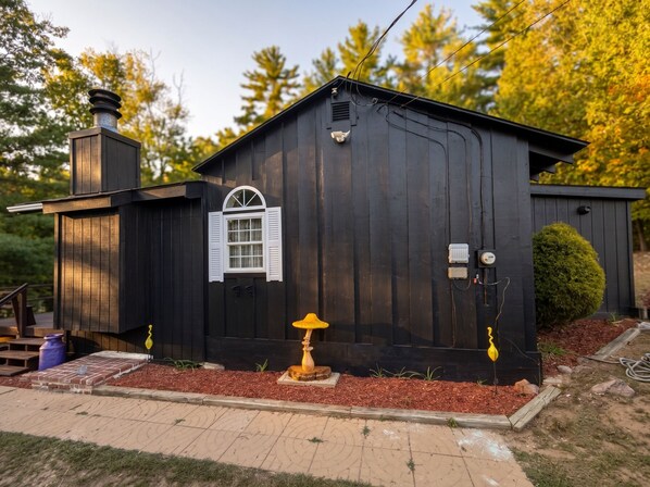 Un bâtiment en bois noir avec une fenêtre blanche, une cheminée et un petit jardin agrémenté de paillis et d’un objet jaune.