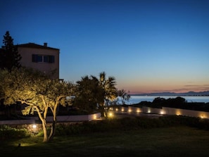 Una casa moderna con un jardín iluminado y vistas al mar al anochecer.