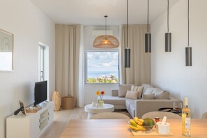 Ein modernes Wohnzimmer mit einer Couch, einem Fernseher, einem Esstisch mit Obst und einer Flasche sowie einem Fenster mit Blick.