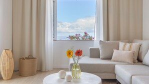 Un salon avec un canapé, une table ronde ornée d’un vase de fleurs, un panier en osier et une fenêtre donnant sur un beau paysage.