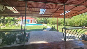 A covered outdoor area with a pool, a patio with seating, and a view of a house in the background. - Dragon Majick (Pavones)
