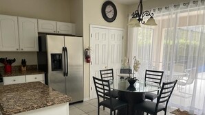 Una cocina con armarios blancos, un refrigerador de acero inoxidable, una mesa de comedor de vidrio con cuatro sillas y un reloj en la pared.