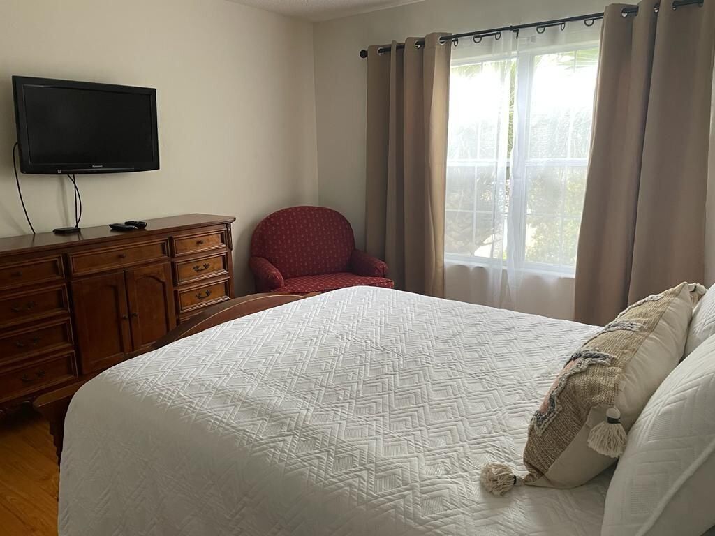 A bedroom with a bed, a television on a wooden dresser, a red chair, and a window with curtains.