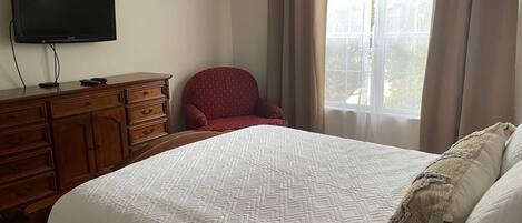 A bedroom with a bed, a television on a wooden dresser, a red chair, and a window with curtains.