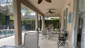 Un patio techado con mesas y sillas de vidrio, un ventilador de techo y vista a una piscina.
