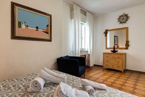 A hotel room with a bed, a chair, a dresser, a painting, and a window with curtains. - Talamone Apartment View by Mamo Florence (Talamone)