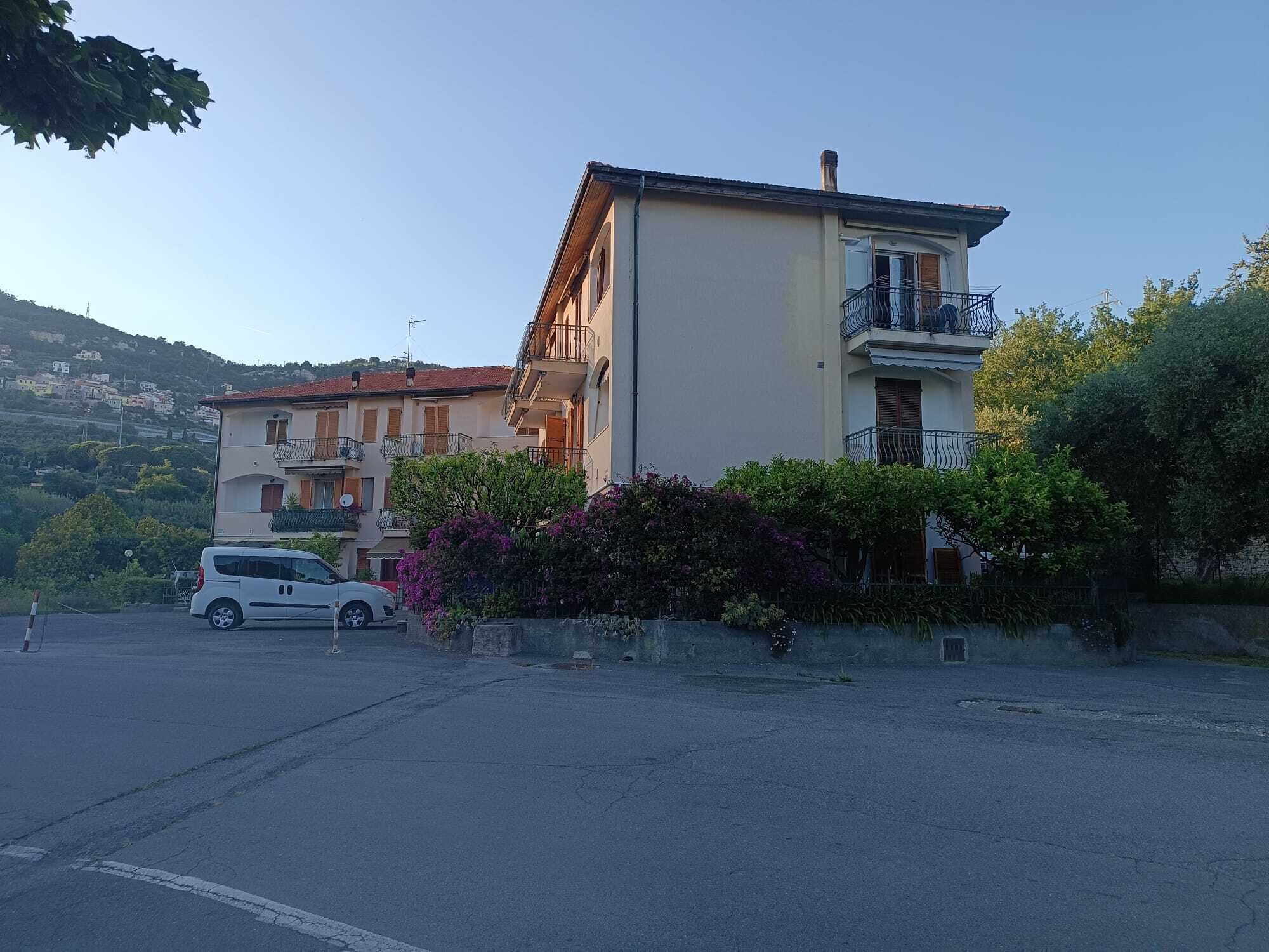 A multi-story building with balconies, a parking lot, and a hilly landscape in the background.